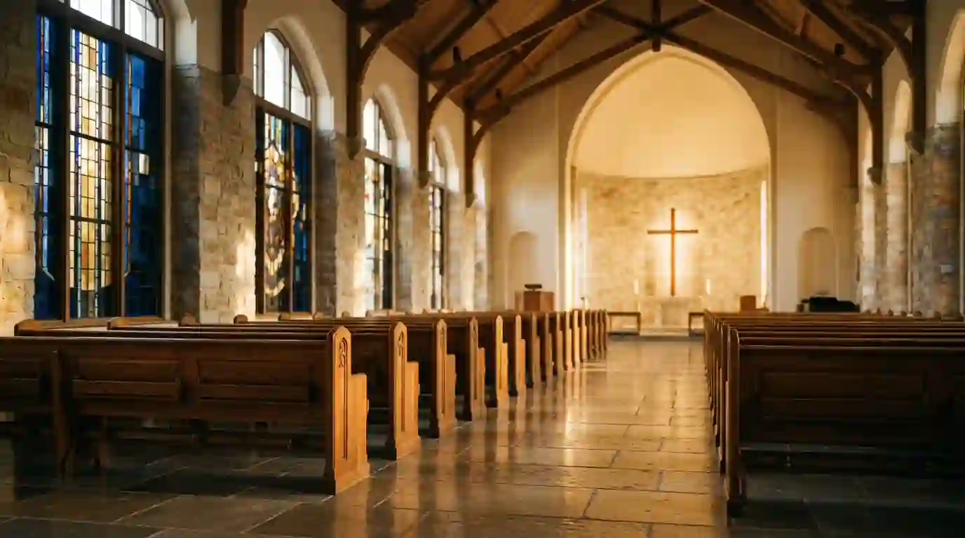 Sunlit interior of a church with stained-glass windows, wooden pews, and a cross at the altar